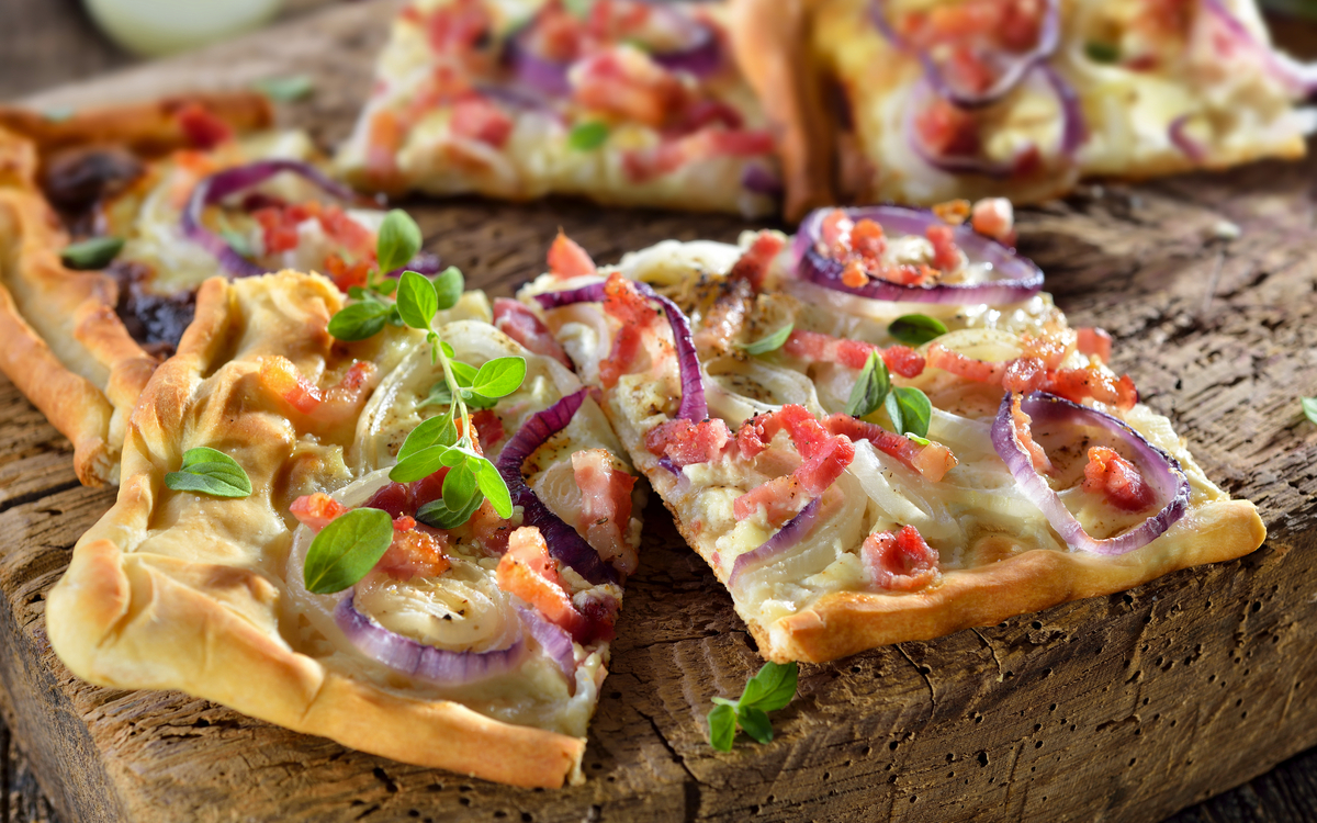 Elsässer Flammkuchen mit Zwiebeln und Speck rustikal auf altem Schneidbrett serviert - Traditional Alsatian tarte flambée  with onions,Speck und saure Sahne serviert auf einem schäbigen Schneidebrett - ©kab-vision - stock.adobe.com