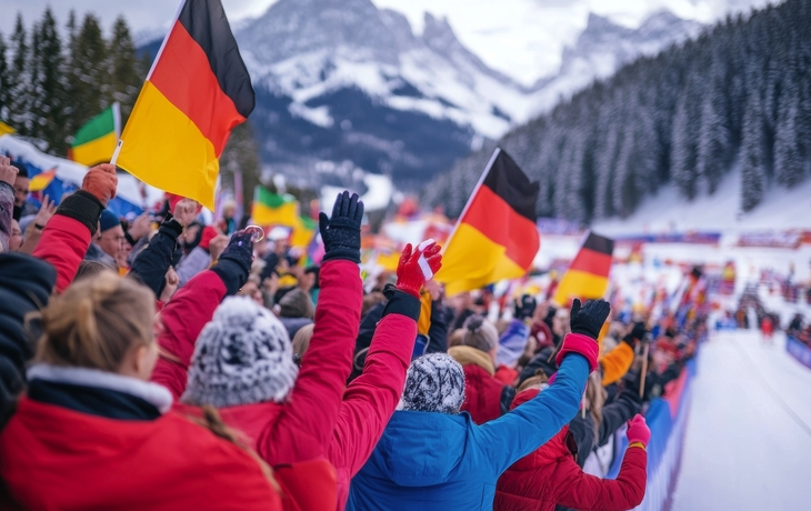 Begeisterte Biathlonfans in Winterjacken, die Deutschlandfahnen schwenken und die Athleten anfeuern, mit verschneiter Biathlonstrecke, Wald und Bergen im Hintergrund. - © master1305 - stock.adobe.com