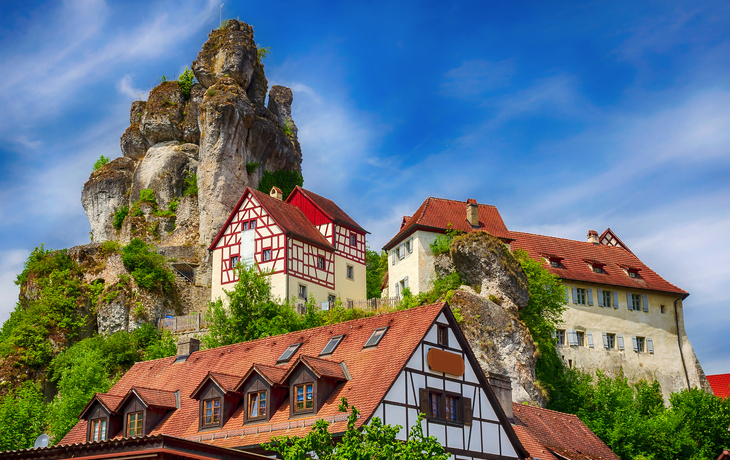 Fachwerkhäuser vor einer felsigen Anhöhe unter blauem Himmel. - © mojolo