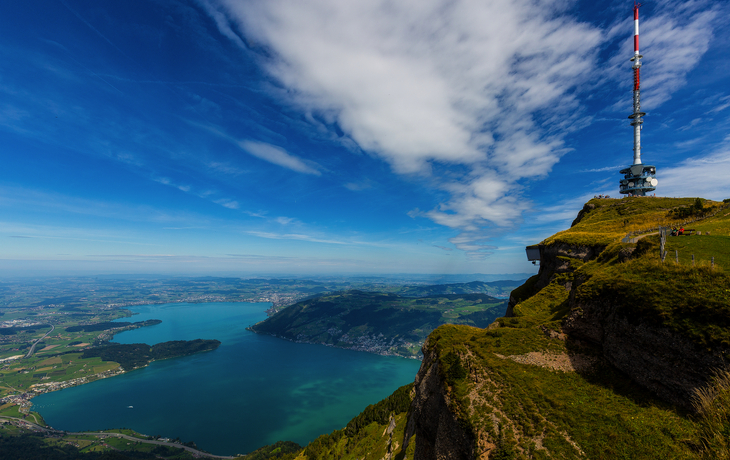 Bergbahn fährt durch blühende Wiese mit Seeblick und Bergen im Hintergrund. - © suteracher - stock.adobe.com